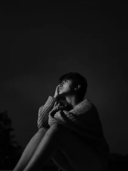 A pensive black and white portrait of a woman sitting against a dark sky, looking upwards. The dramatic, low-key lighting highlights her profile and creates a deeply introspective mood.