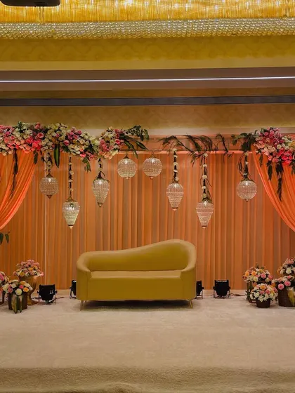 This elegant stage setup doubles as a beautiful photo opportunity. The soft peach drapes, floral arrangements, and hanging glass lamps create a romantic and sophisticated backdrop.