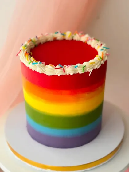 A vibrant rainbow cake with smooth ombre buttercream stripes. The top is finished with a border of piped buttercream and colorful sprinkles.