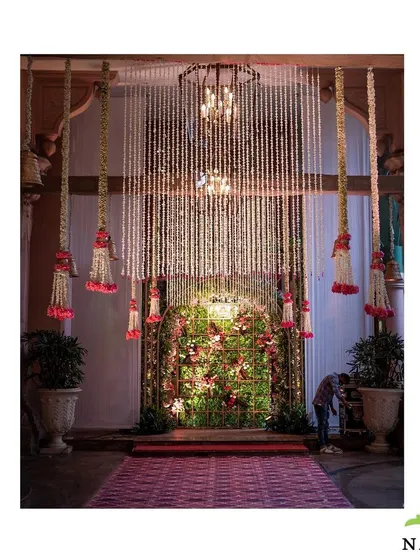 An entrance to a romantic evening event. The archway is covered in lush red and pink roses, with hanging floral strings and chandeliers creating a warm and inviting atmosphere.