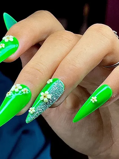 A bright neon green set of stiletto nails, featuring an accent nail with silver glitter and delicate 3D flowers.