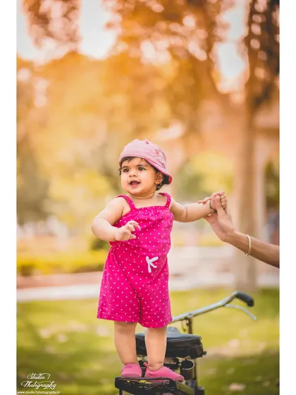 An outdoor first birthday shoot is a wonderful way to capture a baby's new mobility. Here, a little girl in a bright pink outfit stands proudly on a tricycle, holding her parent's hand for support in a beautiful, natural setting.