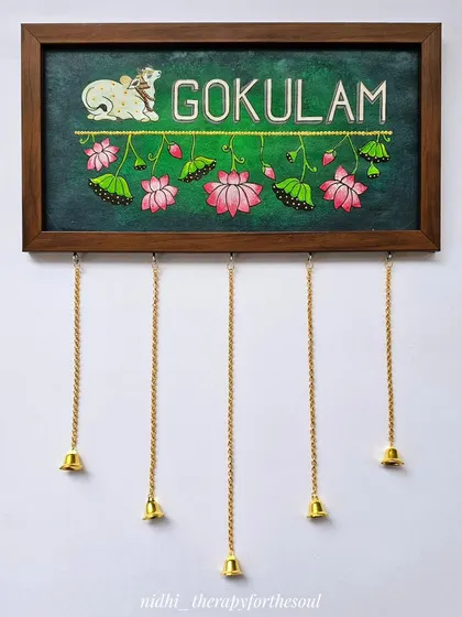 Another view of the 'Gokulam' nameplate, highlighting the intricate details of the Pichwai cow and lotus flowers, complemented by the hanging brass bells.