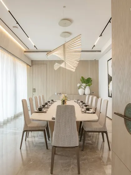 A view from the doorway into the serene dining room, where a long table is set for twelve. The space is defined by its clean lines, neutral palette, and a stunning sculptural light fixture that appears to float above the table.