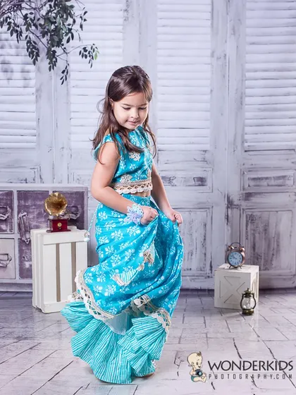 A young girl twirling in a beautiful blue printed lehenga. The motion of the skirt and her happy expression create a dynamic and joyful portrait against a rustic white background.