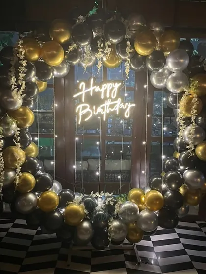 A glamorous birthday balloon ring in black, gold, and silver, set against a window and illuminated by fairy lights.