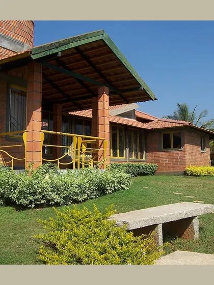 A farmhouse with a welcoming verandah, built with hollow clay blocks and surrounded by a green lawn, embodying relaxed, sustainable living.