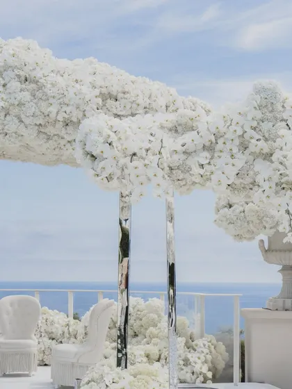 The mandap chairs, upholstered in elegant white, sit beneath a canopy of floral clouds, supported by mirrored pillars that blend seamlessly into the sea and sky.