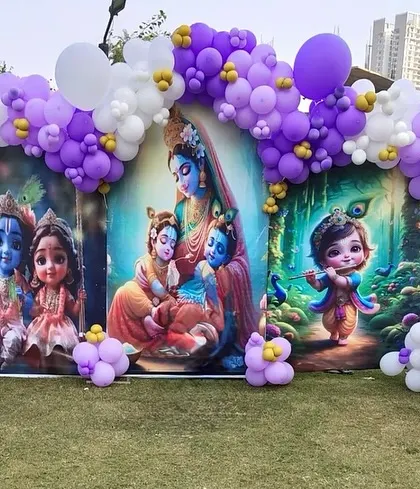 An outdoor Krishna-themed decoration, perfect for a Janmashtami celebration or a themed birthday. The setup includes a large printed backdrop and a purple and white balloon arch.