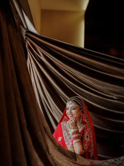 A beautiful portrait of the bride in her red lehenga, peeking out from behind a rich, dark curtain. The play of light and shadow creates a dramatic and intimate feel.