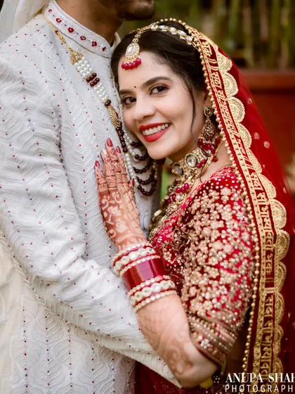 A close-up portrait of the bride, smiling warmly as she embraces her groom. This shot highlights her intricate red lehenga, jewelry, and the beautiful henna on her hands.