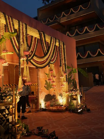 The entrance at dusk, with soft lighting illuminating the floral arch and the traditional decor elements. The scene is warm, inviting, and full of festive promise.