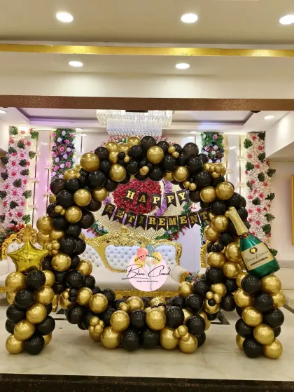 An elegant and grand retirement party decoration in a banquet hall. The setup features a large circular arch with classic black and gold balloons, a 'Happy Retirement' banner, and a champagne bottle foil balloon for a celebratory touch.