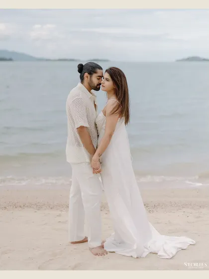 A romantic, full-length portrait of a couple standing by the water's edge on a serene beach in Thailand.