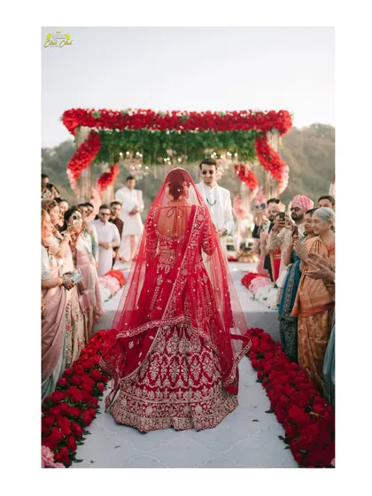 The bride's walk down the aisle. This powerful shot from behind showcases the magnificent red lehenga and the emotional journey towards the groom, with family looking on.
