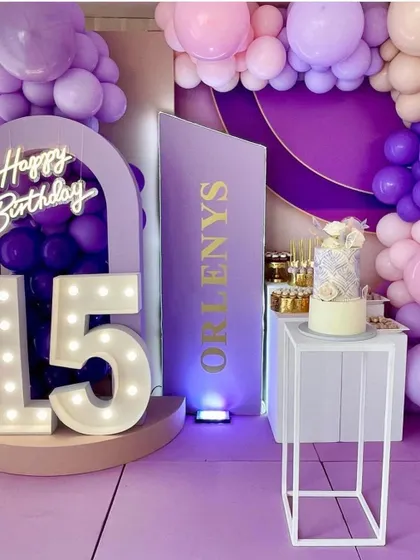 A stylish 15th birthday party with a purple and lavender color scheme. The setup includes marquee numbers, a custom name sign, and a modern cake display.