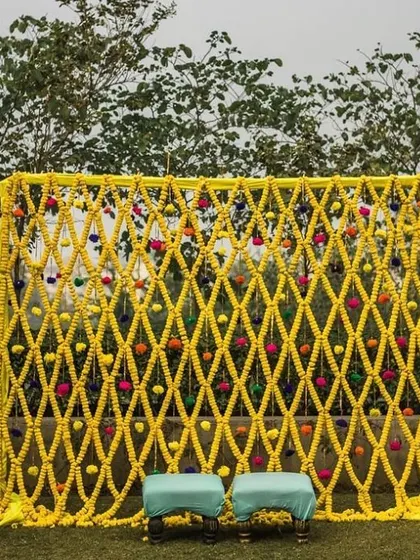 This is a simple yet beautiful photo backdrop for an outdoor haldi event. I created a net of yellow flower strings, dotted with colorful pom poms, which looks lovely against the natural green background.