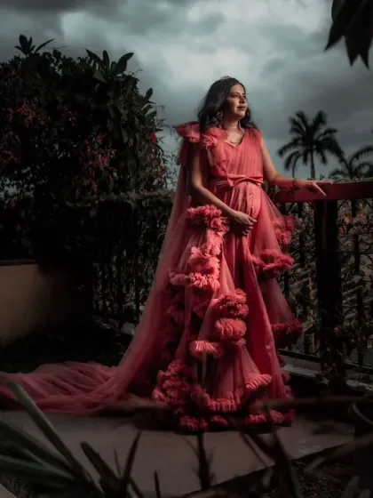A dramatic, moody shot of a client in a pink ruffled gown. The layers of tulle and the flowing cape create a stunning silhouette against the dark, cloudy sky.