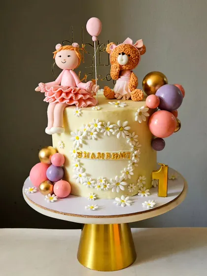 An incredibly detailed cake for a first birthday. This design is loaded with charm, featuring a handcrafted girl figurine, a teddy bear, a daisy wreath, and colorful balloon accents.