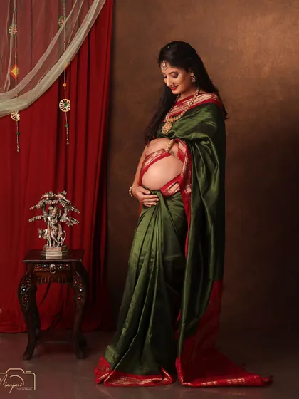 A beautiful solo portrait in a green and red saree, her hand lovingly on her bump. The traditional setup is rich and elegant.