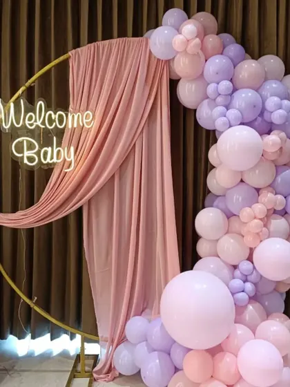 An elegant "Welcome Baby" setup with a draped ring frame and a pastel pink and purple balloon arrangement.
