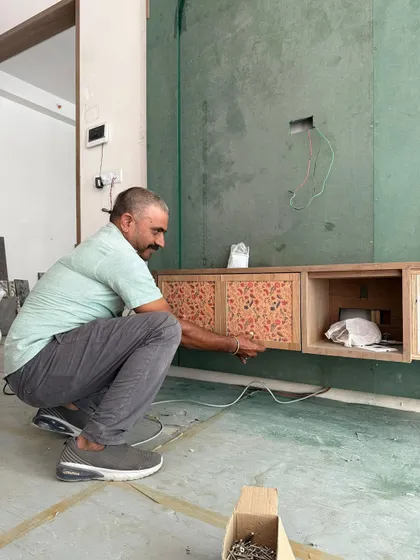 Dust, drills, and design dreams. A carpenter works on installing a custom media unit with unique patterned shutters. This is the 'trust the process' phase of the project.