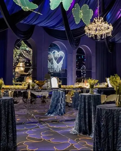 The bar area for the blue cocktail event, set against a backdrop of elegant arches and a neon sign of two swans. The mirrored bar front reflects the deep colors and lights of the room.