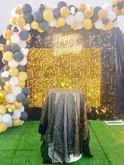 An outdoor birthday setup with a gold and black theme. The decor features a gold shimmer wall, a black and gold balloon arch, and a sequined cake table.