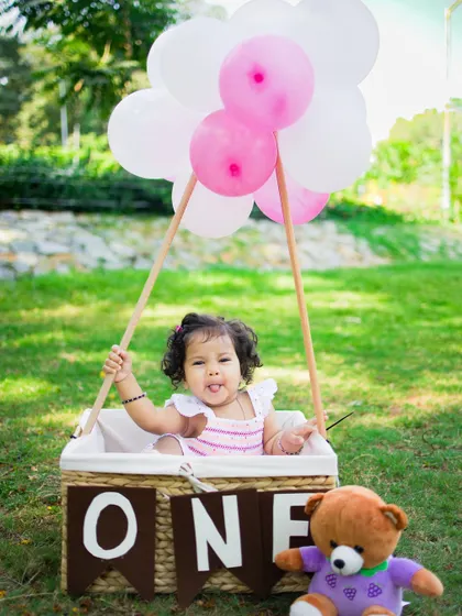 An outdoor first birthday shoot with a hot air balloon prop. Her playful expression and the beautiful park setting make this a truly joyful and memorable portrait.