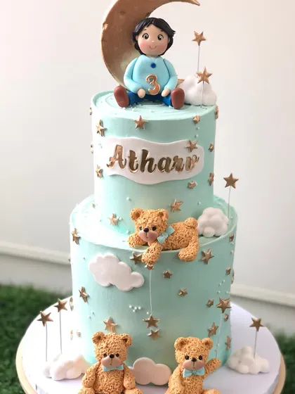 A dreamy two-tiered cake with a "boy on the moon" theme, surrounded by teddy bears and golden stars.