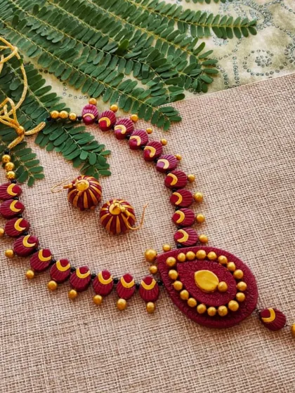 A beautiful teardrop-shaped necklace in maroon and gold with intricate detailing. The set includes a cute matching hanging jhumka.