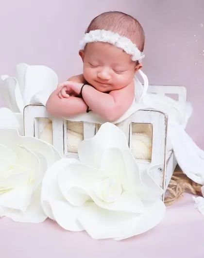 A vision in white. This baby is posed on a miniature bed surrounded by large, elegant paper flowers, creating a sophisticated and angelic scene.