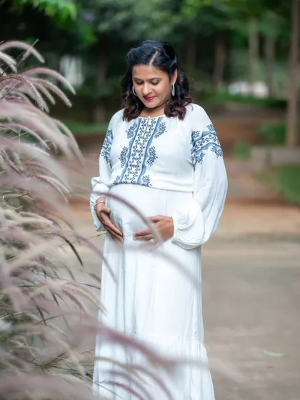 A serene outdoor portrait of a mom-to-be in a white embroidered dress, cradling her bump amidst tall, soft grasses.
