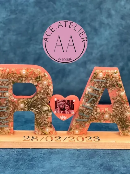 A unique take on varmala preservation. The couple's initials, 'R' and 'A', are created as large resin letters filled with their wedding flowers, connected by a heart with their photo, and set on a personalized wooden base.