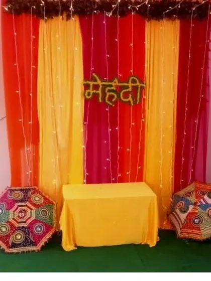 A simple Mehendi backdrop for a home function, using orange and yellow drapes, fairy lights, and decorative umbrellas.