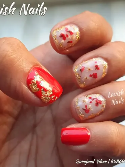 A close-up of nail art on short, natural nails, featuring a red base with gold foil and accent nails with suspended red petals.