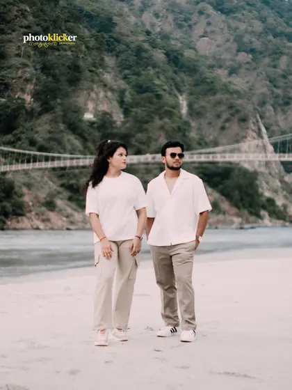A full-body shot of a couple standing on a riverbank in Rishikesh, holding hands with a suspension bridge in the background.