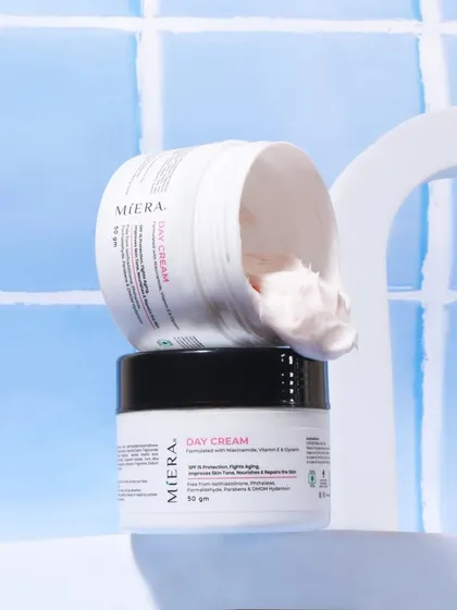 A creative and abstract shot of a day cream, with the lid off and the cream showing. The product is balanced on itself against a water color tile background, creating an artistic and clean image.
