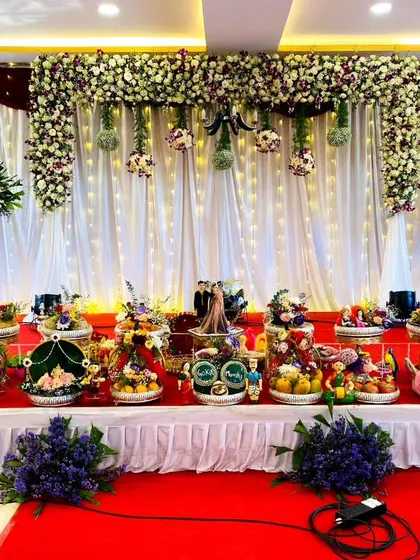 A classic and elegant stage design for a wedding or engagement. The backdrop features white drapes with floral accents and hanging arrangements, with a red carpet leading to a table filled with my custom ceremonial trays.