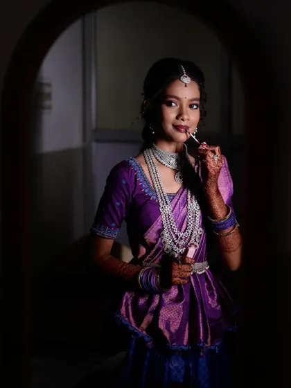 A candid "getting ready" shot from a fashion-style bridal shoot. The bride is touching up her lipstick, captured through an archway for a creative composition.