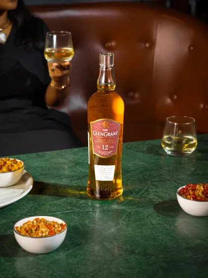 A lifestyle shot for The Glen Grant, capturing a moment of enjoyment. The composition places the product at the center of a social setting, using a shallow depth of field to draw focus to the bottle while hinting at the environment.
