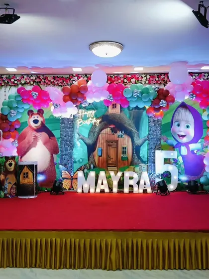 A vibrant Masha and the Bear theme for a 5th birthday. The stage is filled with character cutouts from the show, set against a forest backdrop and framed by a colorful balloon arrangement.