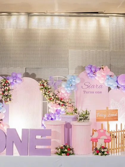 A whimsical fairy-themed first birthday setup in pastel purples and pinks, featuring paper flowers, mushroom props, and a large marquee "ONE."