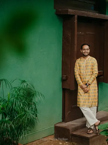 The groom, in a yellow kurta, stands by a doorway, the green foliage and natural light creating a fresh and vibrant portrait.