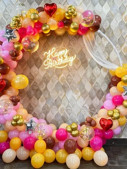 A vibrant and colorful birthday balloon ring in shades of pink, yellow, and brown, with a "Happy Birthday" neon sign.
