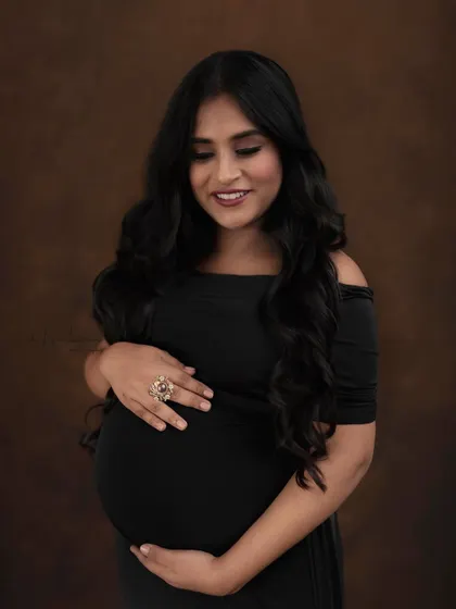 A close-up portrait against a warm, brown backdrop. The focus is on her beautiful, glowing smile and the simple elegance of the off-shoulder black dress.