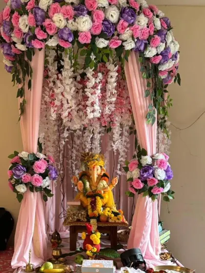 A lovely pastel-themed Ganpati mandap with pink, purple, and white flowers creating a canopy. The hanging wisteria and soft pink drapes add to the dreamy and gentle aesthetic.