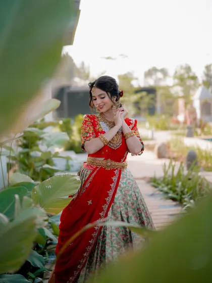 A creative shot through foliage adds depth and a sense of discovery. The bride looks radiant in her red and green lehenga, framed by nature.