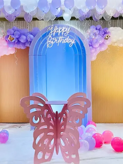 A beautiful and clean butterfly-themed birthday setup. The decor features a central blue lighted arch against a neutral backdrop, framed by pastel balloons and a delicate butterfly prop.