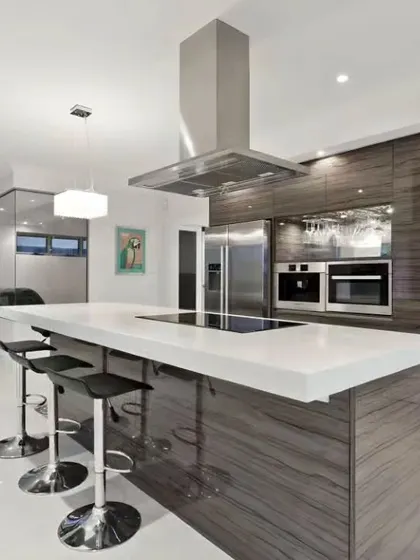 A luxury kitchen centered around a large island with an integrated cooktop and seating. The high-gloss wood-grain finish and stainless steel chimney hood add to the premium feel.